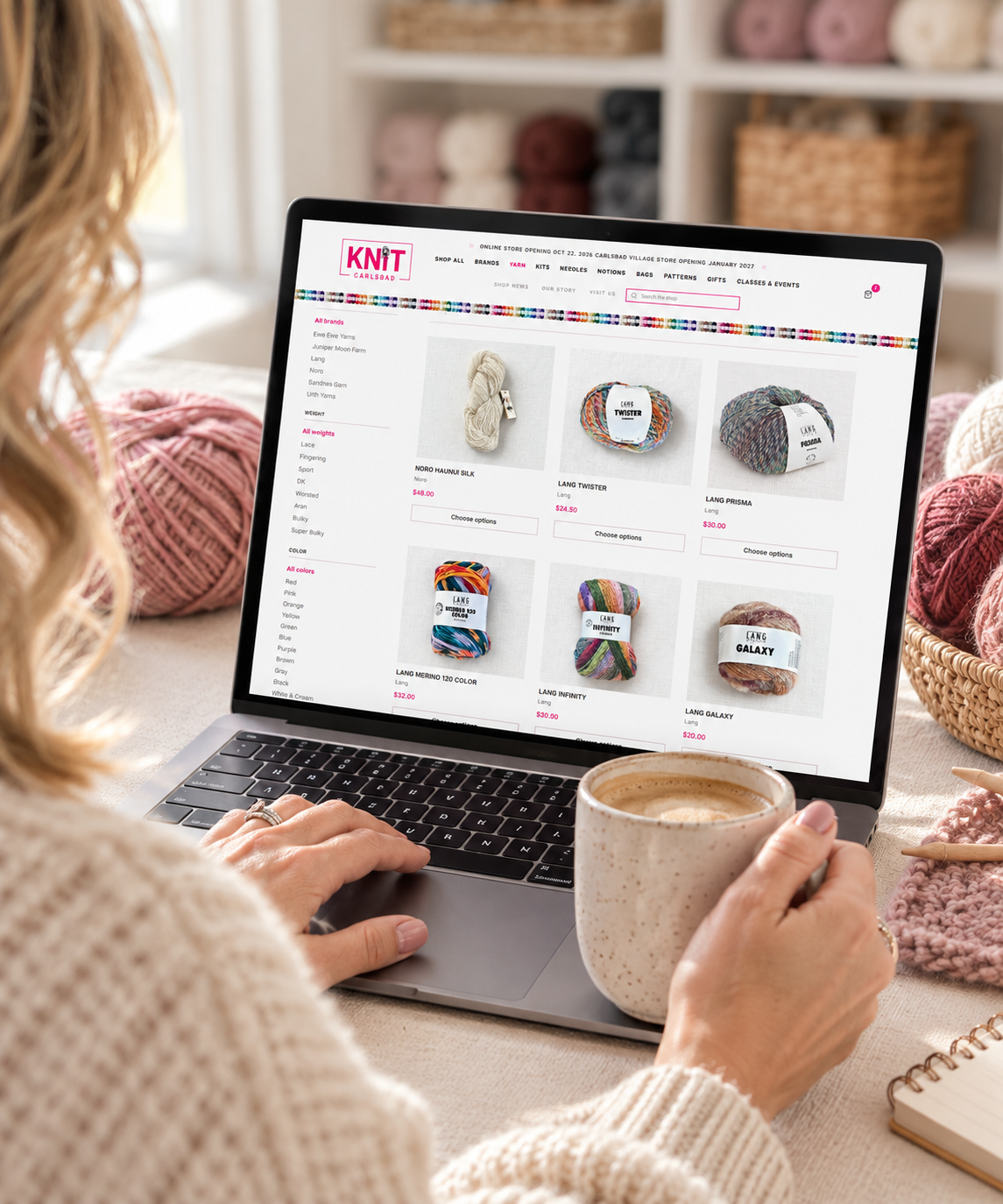 A person in a cream cable-knit sweater typing on a laptop showing the Knit Carlsbad online store, with pink yarn and knitting supplies on the table