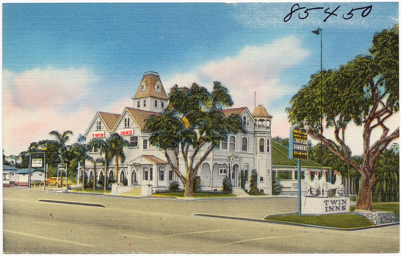 A vintage postcard of the historic Twin Inns, a white Victorian building with a tower, palm trees, and chicken statues by the sign
