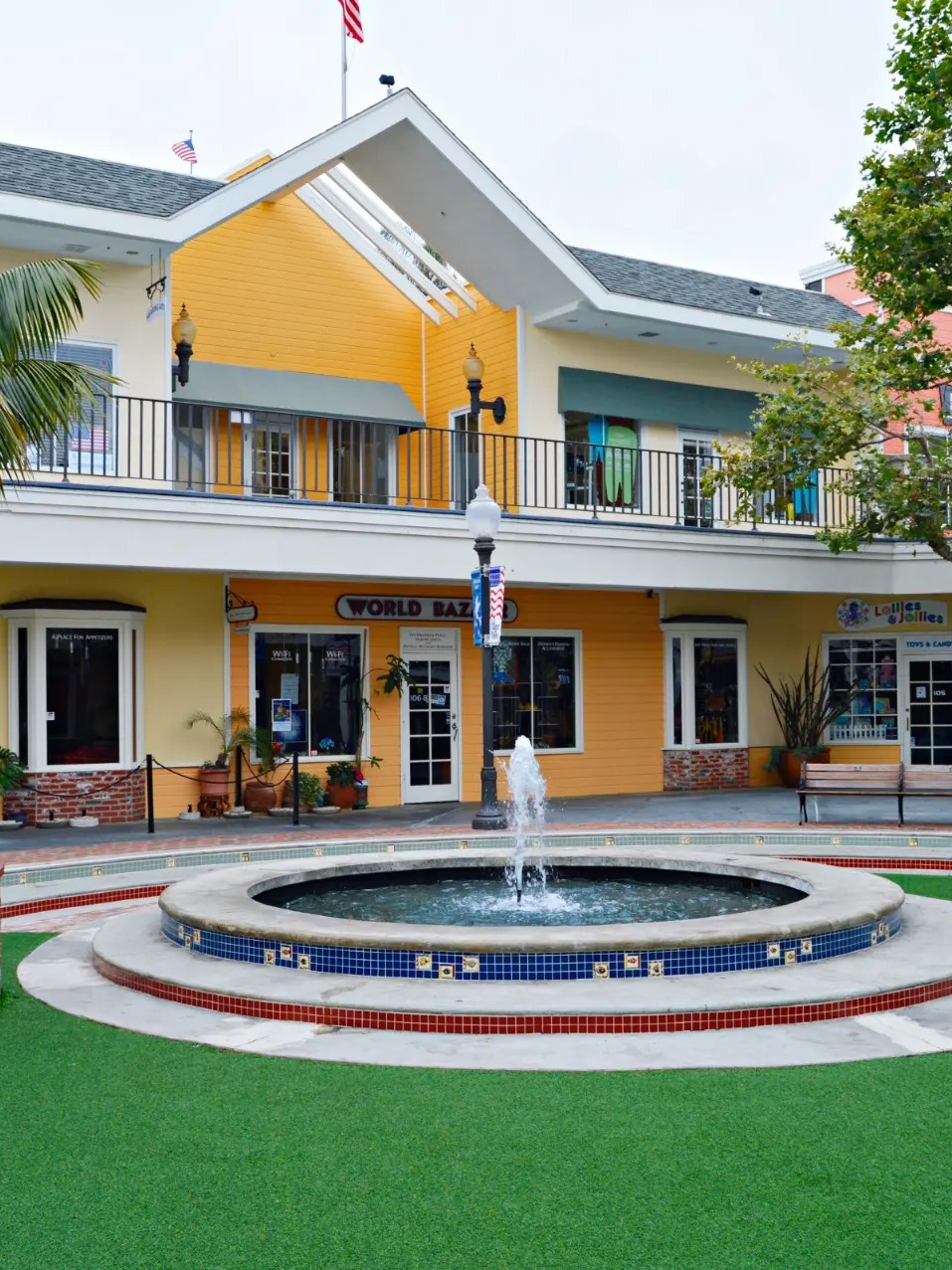 The Village Faire courtyard with a tiered water fountain, brick flower planters, palm trees, and two-story shops